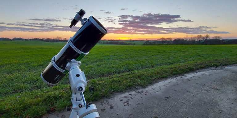 Ein Teleskop steht vor einem Feld in der freien Natur. Im Hintergrund ist der Rest vom Sonnenuntergang zu sehen.