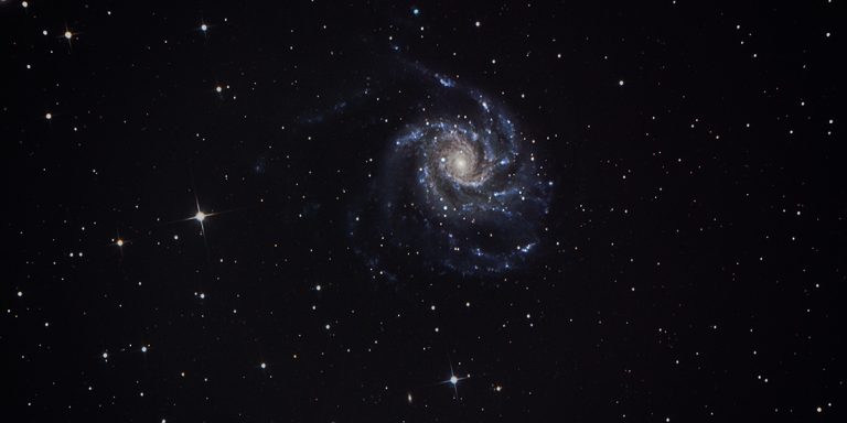 Die Pinwheel-Galaxie, eine Spiralgalaxie, auf die man von oben sieht. Man sieht deutlich die gelbhelle Mitte und die blau-violetten Spiralarme drumherum.
