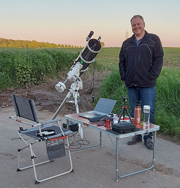 Ein Foto von mir, wie ich neben meinem Teleskop am Feldrand stehe. Vor mir ein Campingtisch mit Stuhl.